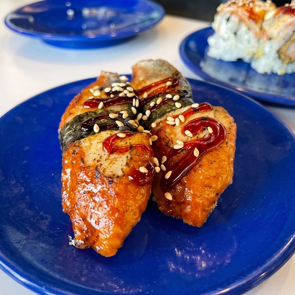 Glazed grilled salmon pieces topped with sesame seeds on a blue plate, with sushi rolls on another plate behind.