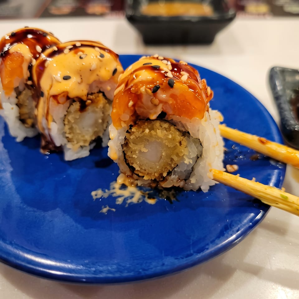 Sushi rolls topped with glazed salmon and sesame seeds on a blue plate, with chopsticks and soy sauce nearby.
