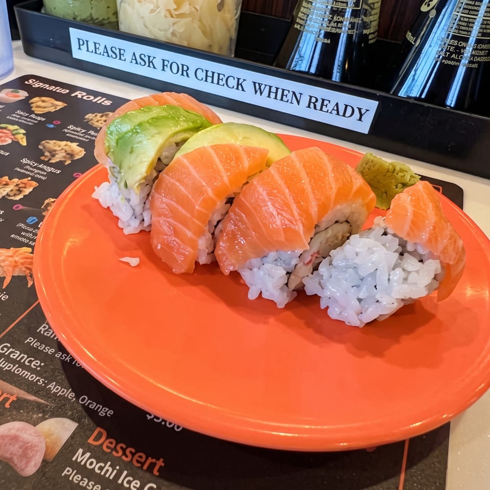 Orange plate of assorted sushi, including salmon nigiri and avocado-topped rolls, beside a restaurant menu and check sign.