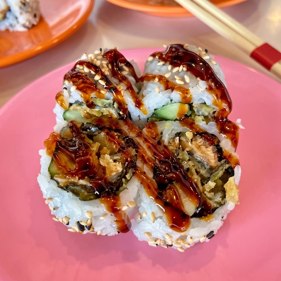 Sushi rolls topped with sweet dark sauce and sesame seeds on a bright pink plate, with chopsticks in the background.