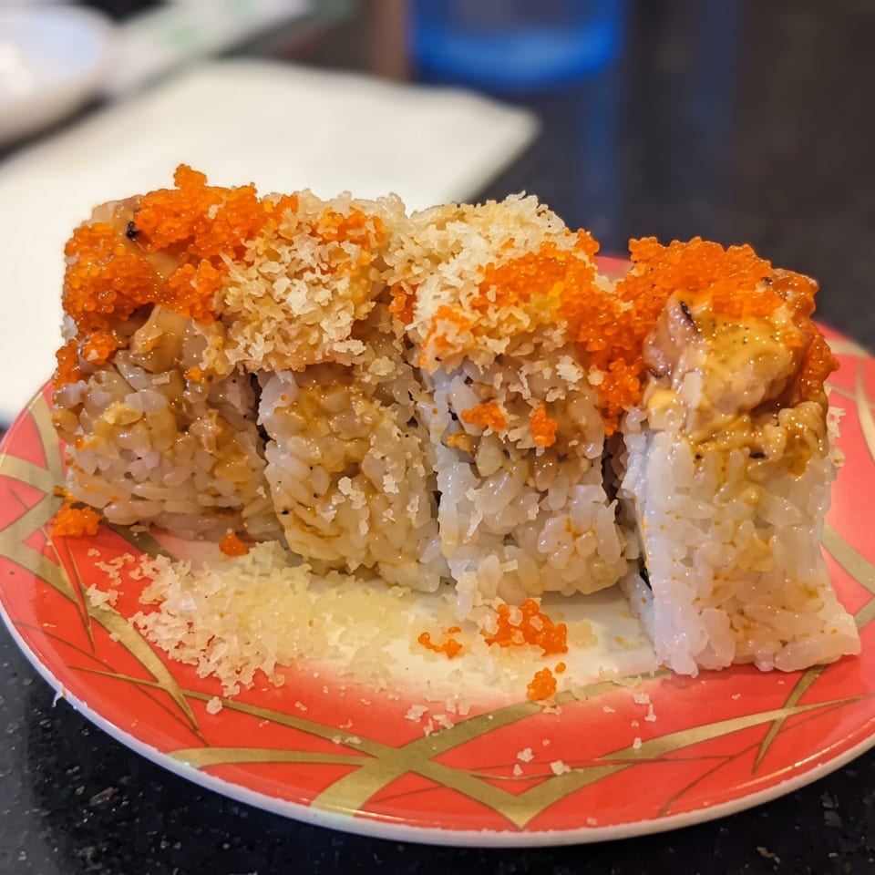 Close-up of sushi rolls topped with orange roe, creamy sauce, and crispy tempura flakes on a red patterned plate.