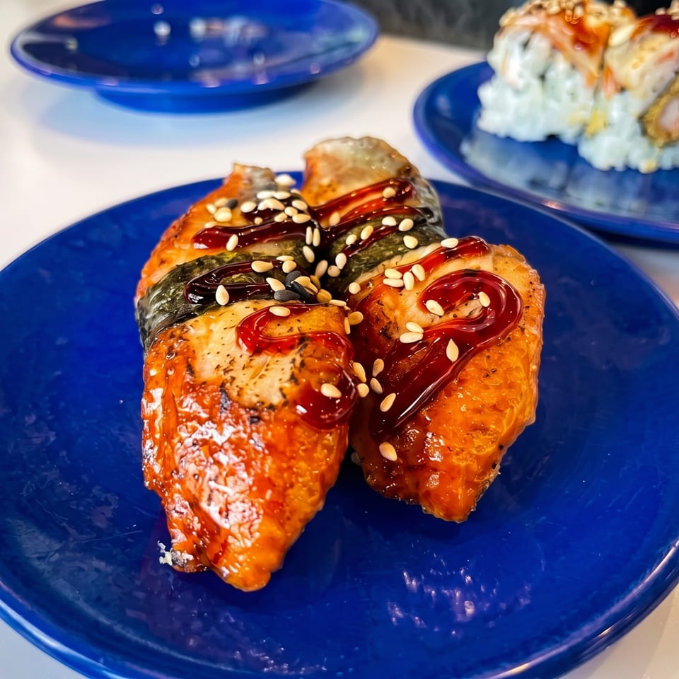 Glazed salmon fillet topped with sesame seeds on a blue plate, with sushi rolls on another plate in the background.