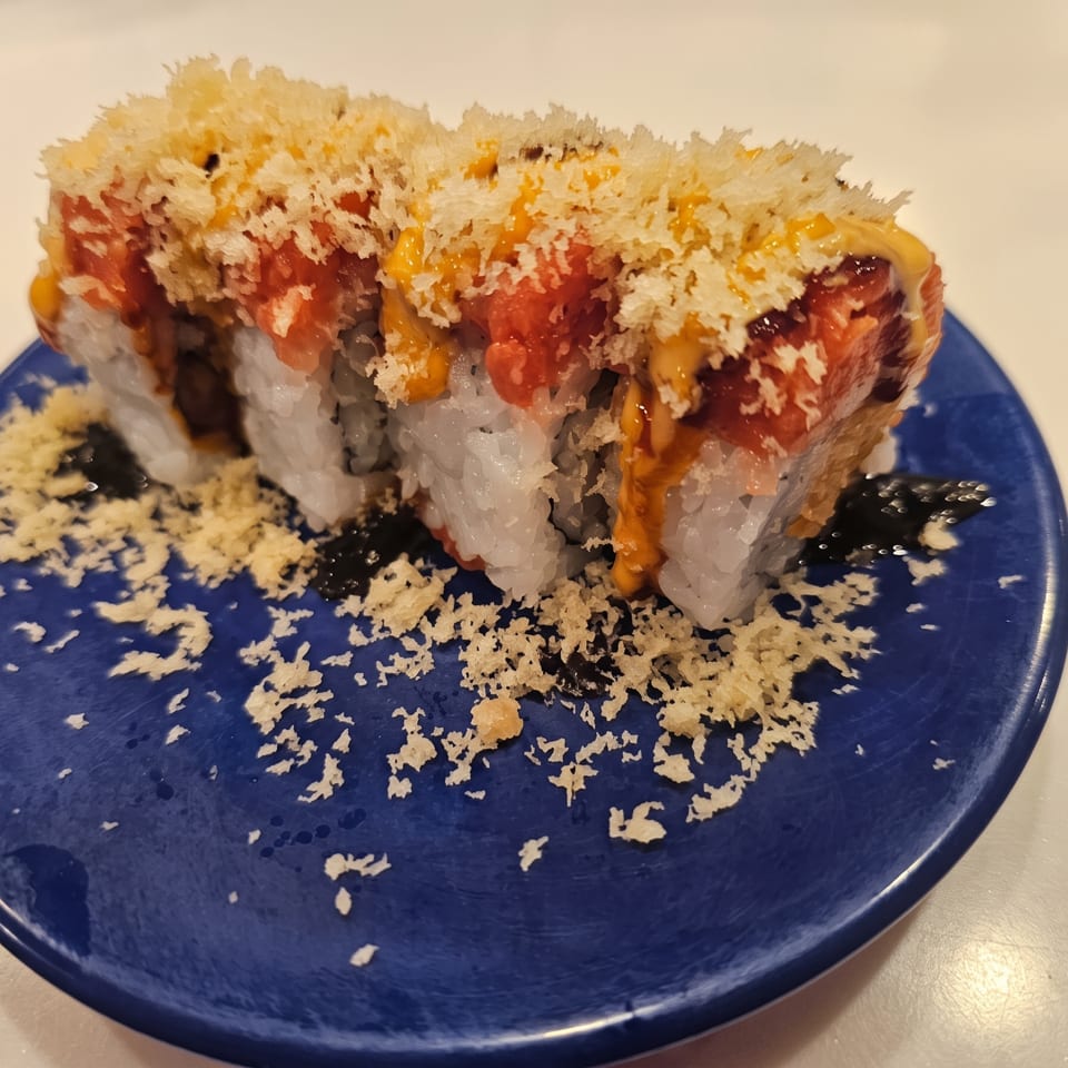 Sushi roll topped with crispy tempura crumbs, salmon, and orange sauce on a blue plate.