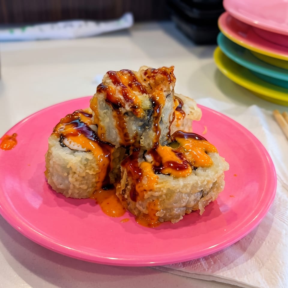 Crispy sushi rolls drizzled with orange and dark sauces on a bright pink plate, with colorful plates and chopsticks nearby.